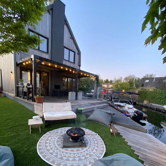 Moderne Douglas pergola met zwart gepoedercoate koppelstukken, bevestigd aan een gevel. De overkapping biedt sfeervolle verlichting en een stijlvolle uitbreiding van het terras. Perfect voor een luxe buitenruimte met bescherming tegen zon en regen. Geïntegreerd in een moderne tuin aan het water